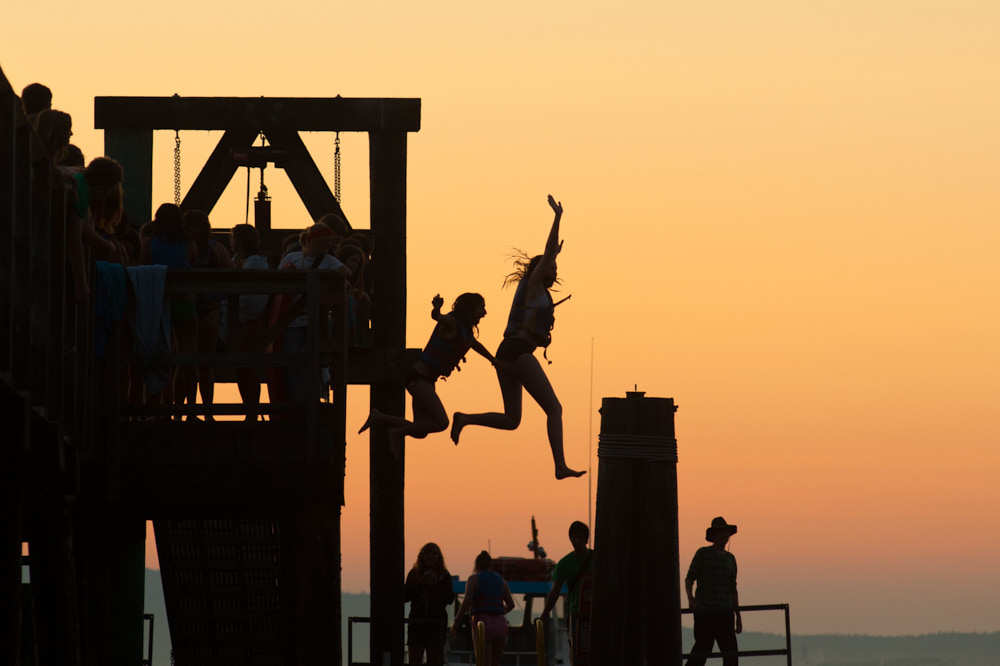 Silhouetted children jump off a dock at sunset