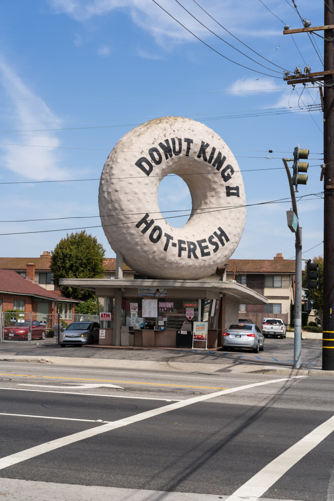 Cars driving past the immense Donut King II building, which is shaped like a donut.