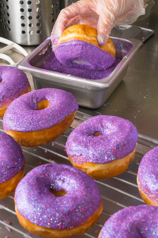 A hand applying bright purple glitter to donuts