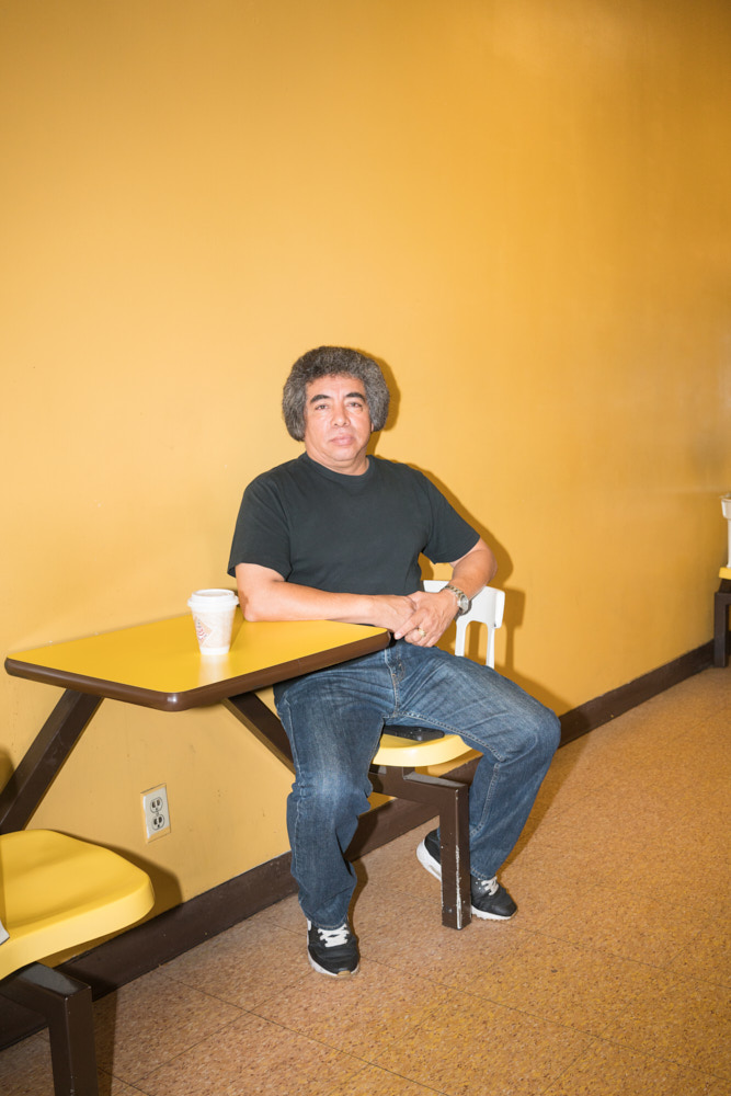 A man with big hair sitting next to a yellow wall with his coffee