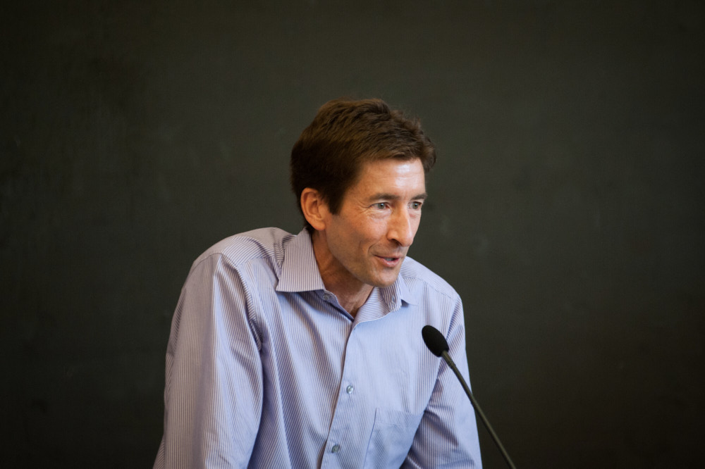 A professor at a microphone with a chalkboard behind him