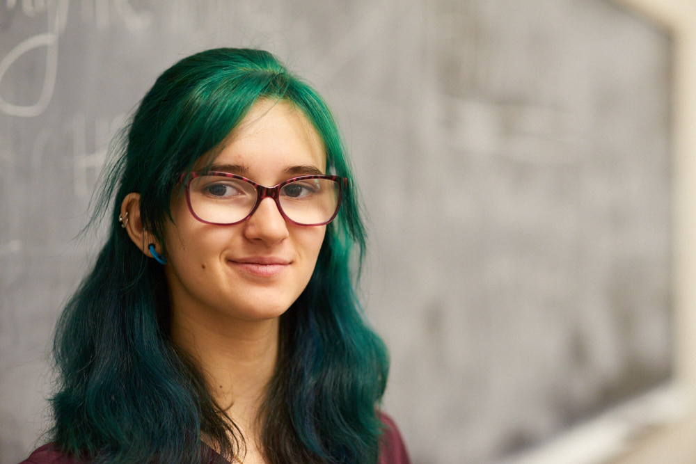 A female student with long green hair poses for a portrait in front of a chalkboard